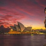 : A smiling Indian female tourist with a backpack posing in front of the illuminated Sydney Opera House at sunset, representing the ultimate Australia Tourist Visa for Indians step-by-step guide.